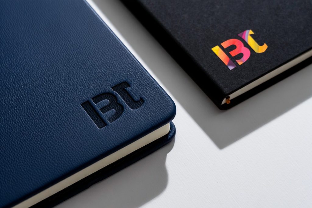 Macro comparison of a blind debossed logo on a navy blue leather notebook versus a vibrant full-color direct digital printed logo on a black hardcover journal.